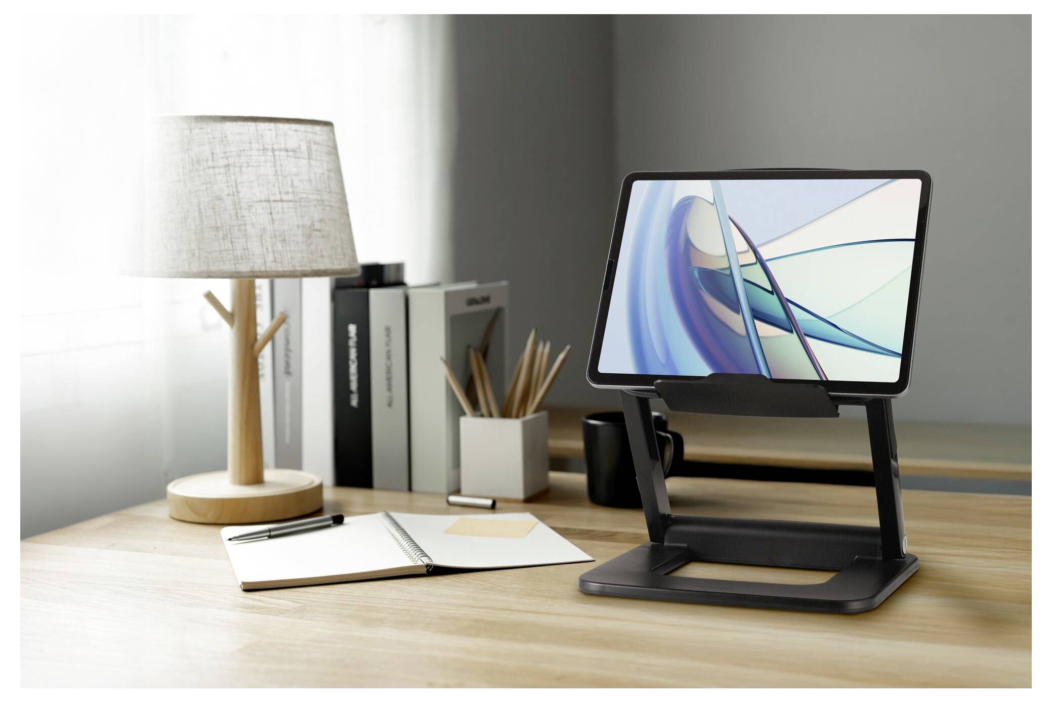 Nowoczesne biuro z tabletem na stojaku, notatnikiem, długopisem i lampą na drewnianym biurku, obok okna.