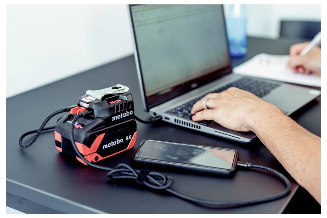 Osoba korzystająca z laptopa przy biurku, z telefonem komórkowym i ładowarką do akumulatora elektronarzędzia w pobliżu.