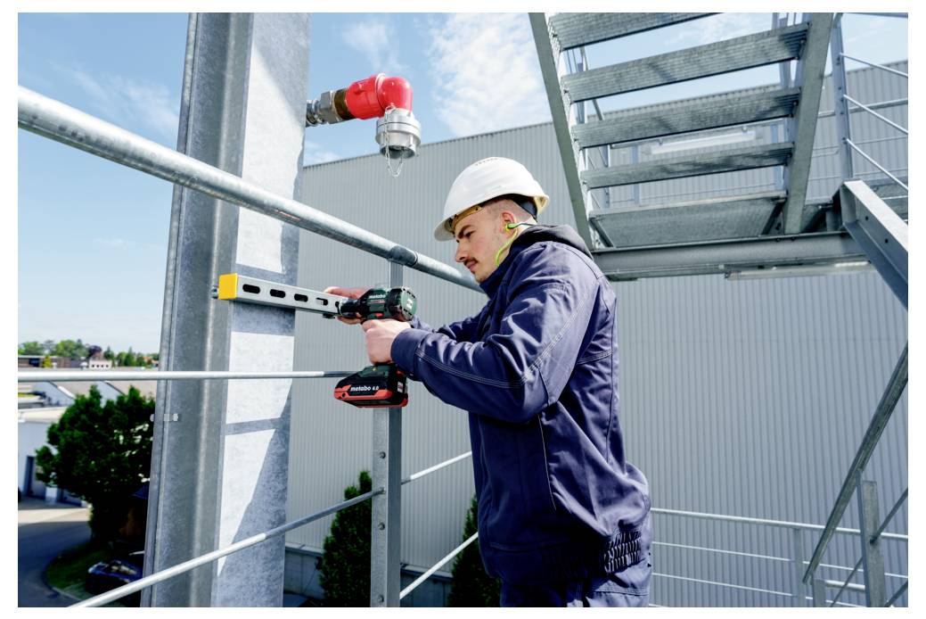 Robotnik budowlany w białym kasku ochronnym i niebieskim uniformie używa wiertarki akumulatorowej do zamocowania belki na zewnątrz, w pobliżu metalowych schodów.