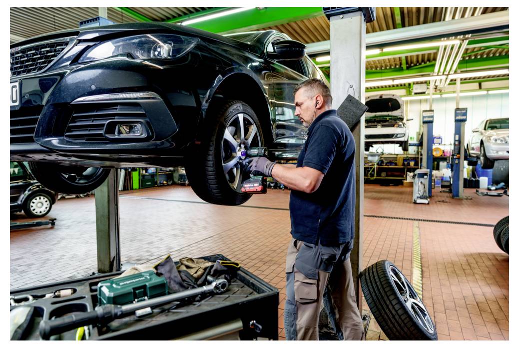 Mechanik w warsztacie kontroluje podniesiony czarny samochód, koncentrując się na przednim kole, wokół niego rozłożone są narzędzia i sprzęt.