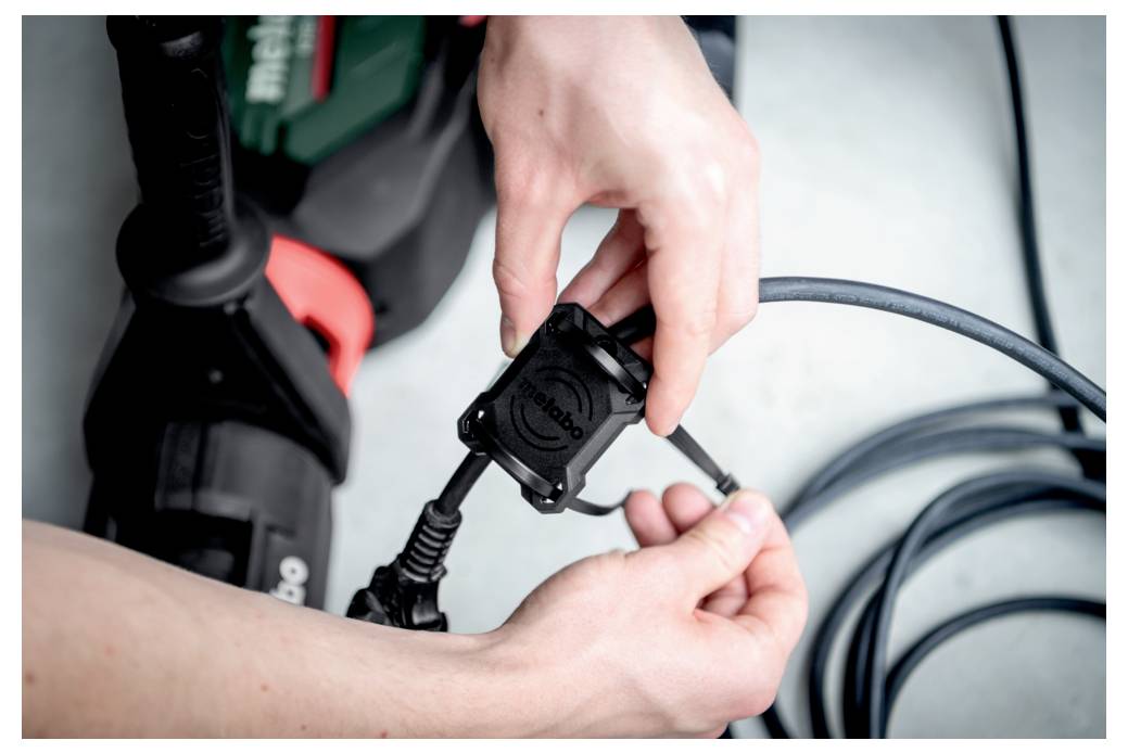 Ręce podłączają wtyczkę elektronarzędzia do gniazda marki Metabo. Narzędzie i przewód są ułożone na jasnej powierzchni.