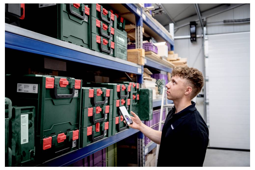 Osoba korzystająca z urządzenia mobilnego do skanowania zielonych pudełek magazynowych z czerwonymi zatrzaskami, znajdujących się na półkach w przestrzeni magazynowej.