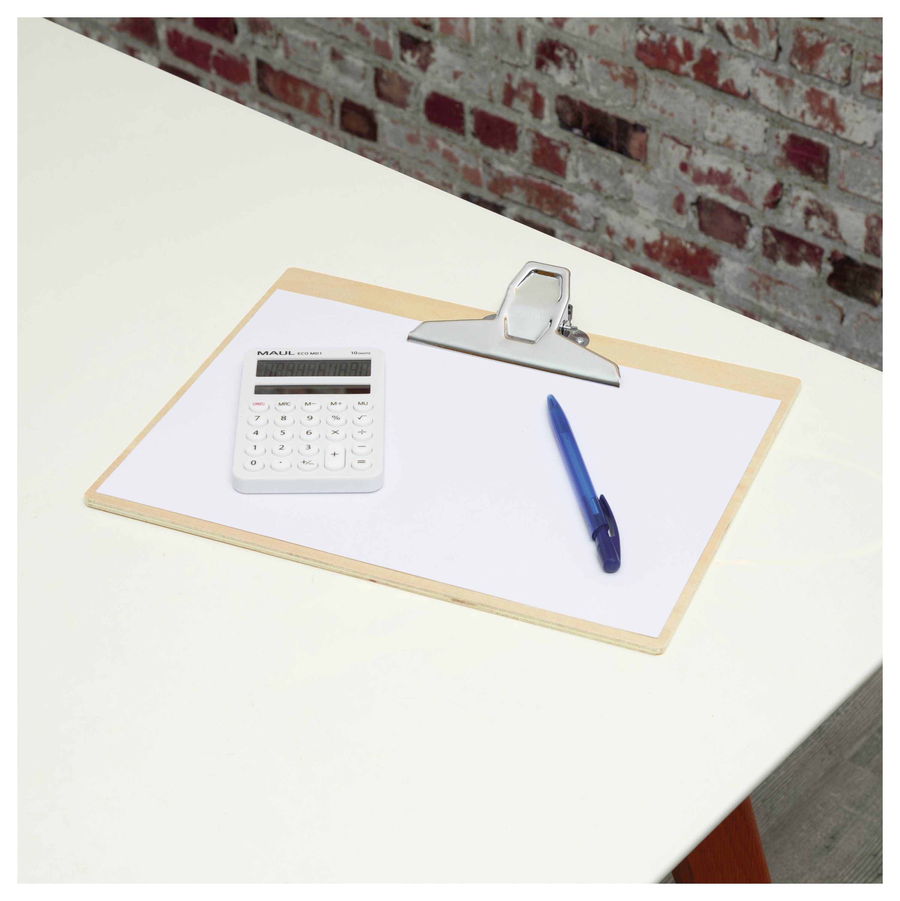 Schowek trzyma pustą kartkę papieru na białym biurku, obok niego leży niebieski długopis i biały kalkulator, na tle ściany z cegły.