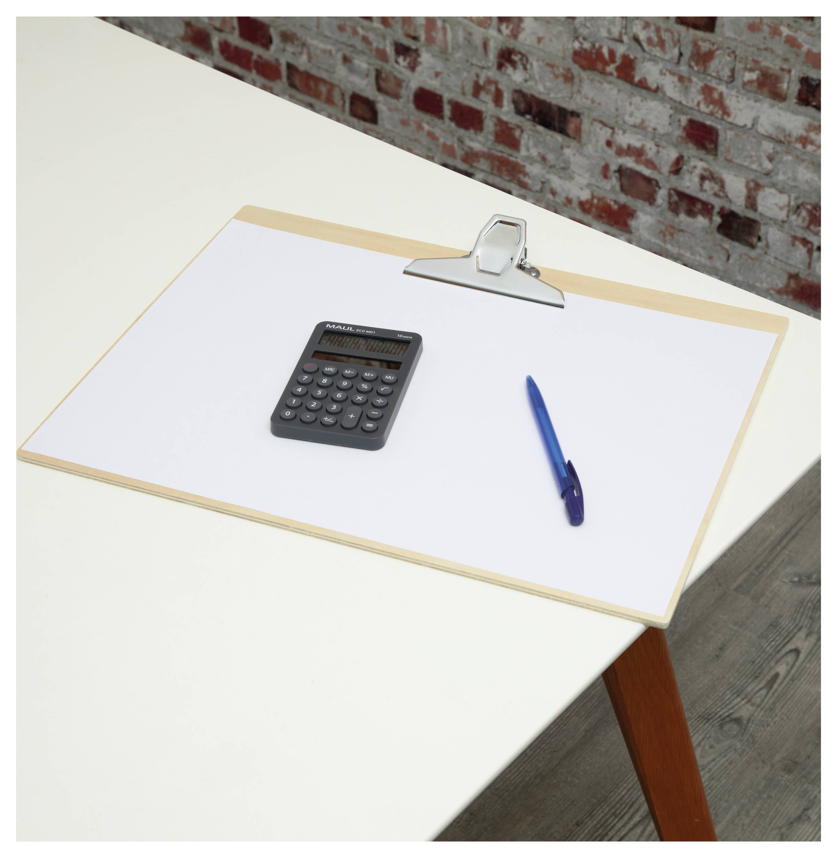Kalkulator, niebieski długopis i clipboard z pustą kartką papieru leżą na białym stole, a w tle widać ścianę z cegły.