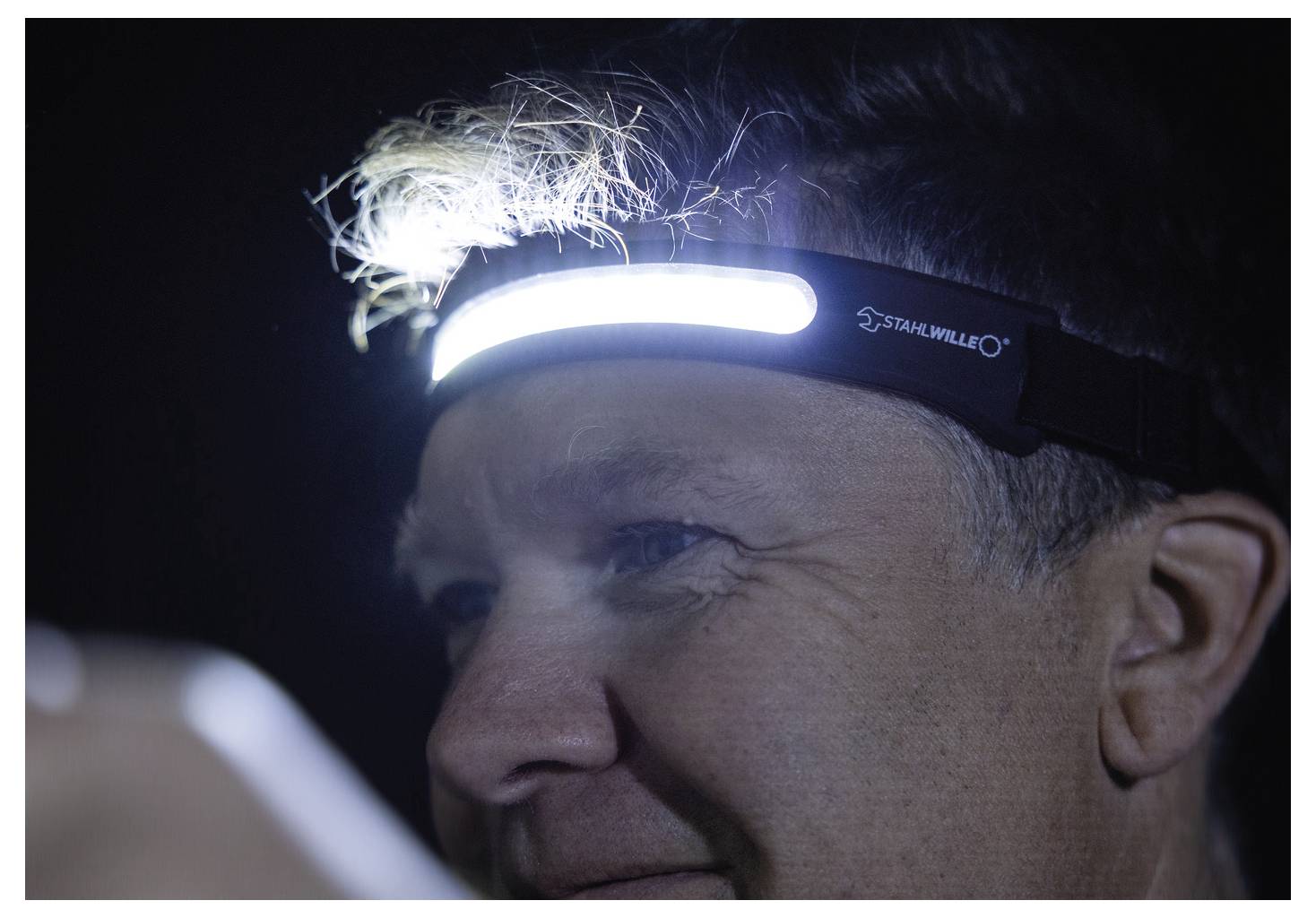 Osoba nosząca czołówkę z jasnym paskiem LED, oświetlająca swoją twarz w ciemnym otoczeniu. Wydaje się, że jest na dworze w nocy.