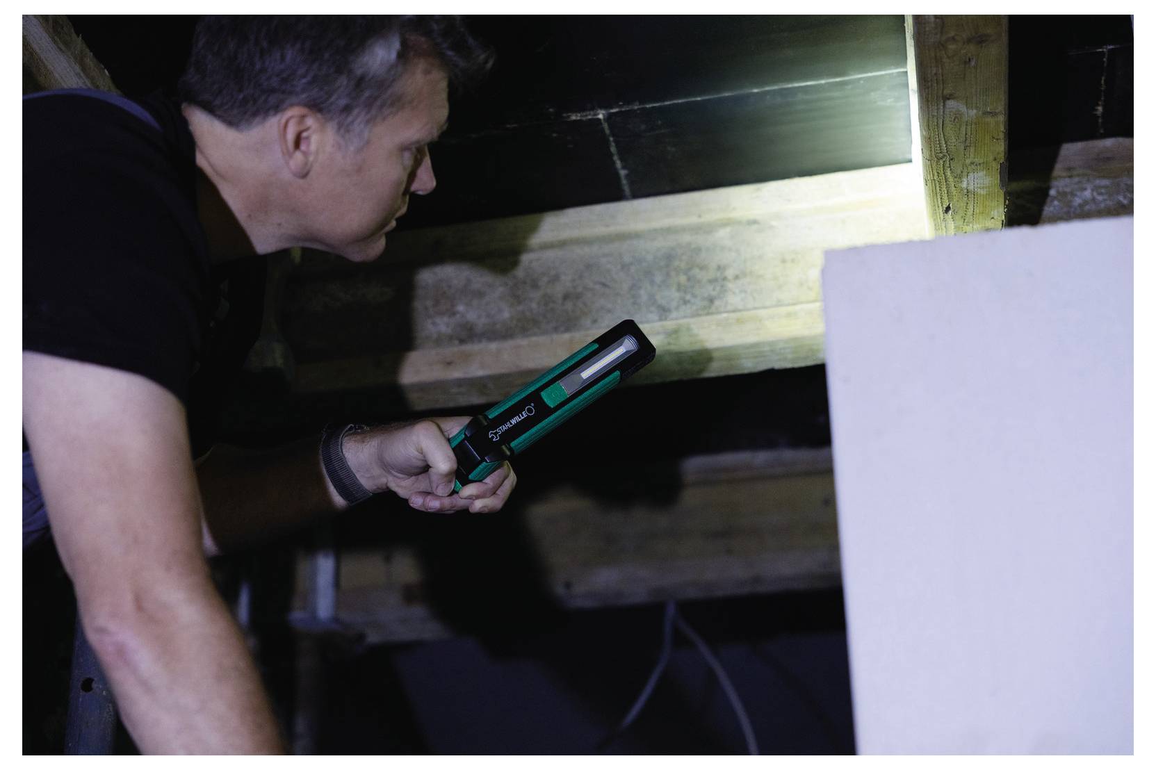 Osoba bada drewniany belk przy świetle latarki w słabo oświetlonym strychu, podświetlając teksturę i strukturę belki.