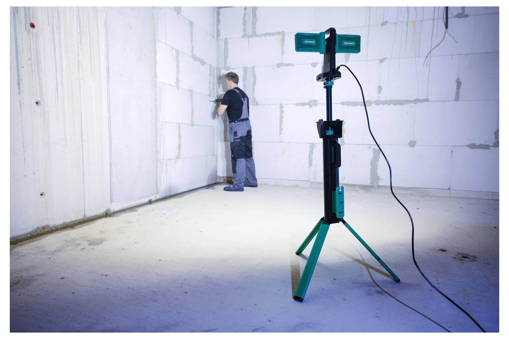 Osoba w stroju roboczym używa narzędzi na białej ścianie w jasno oświetlonym pomieszczeniu, z trójnożnym stojakiem na światło w pierwszym planie.