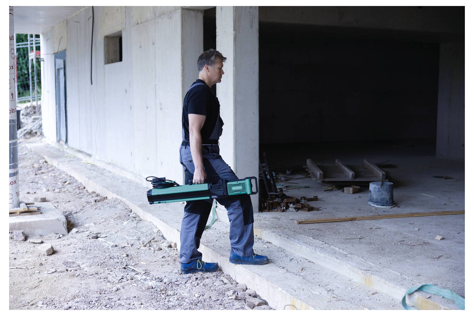 Osoba w stroju roboczym używająca urządzenia do skanowania laserowego na placu budowy. Teren jest częściowo zabudowany, z widocznymi powierzchniami betonowymi.
