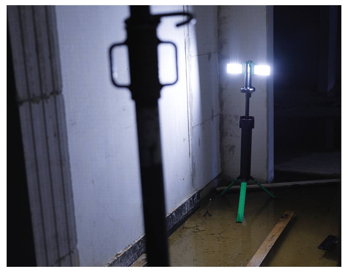 'Zamontowana na statywie lampa robocza oświetla ciemny, częściowo zalany teren budowy, podświetlając niedokończone ściany i rozrzucone drewniane deski.'