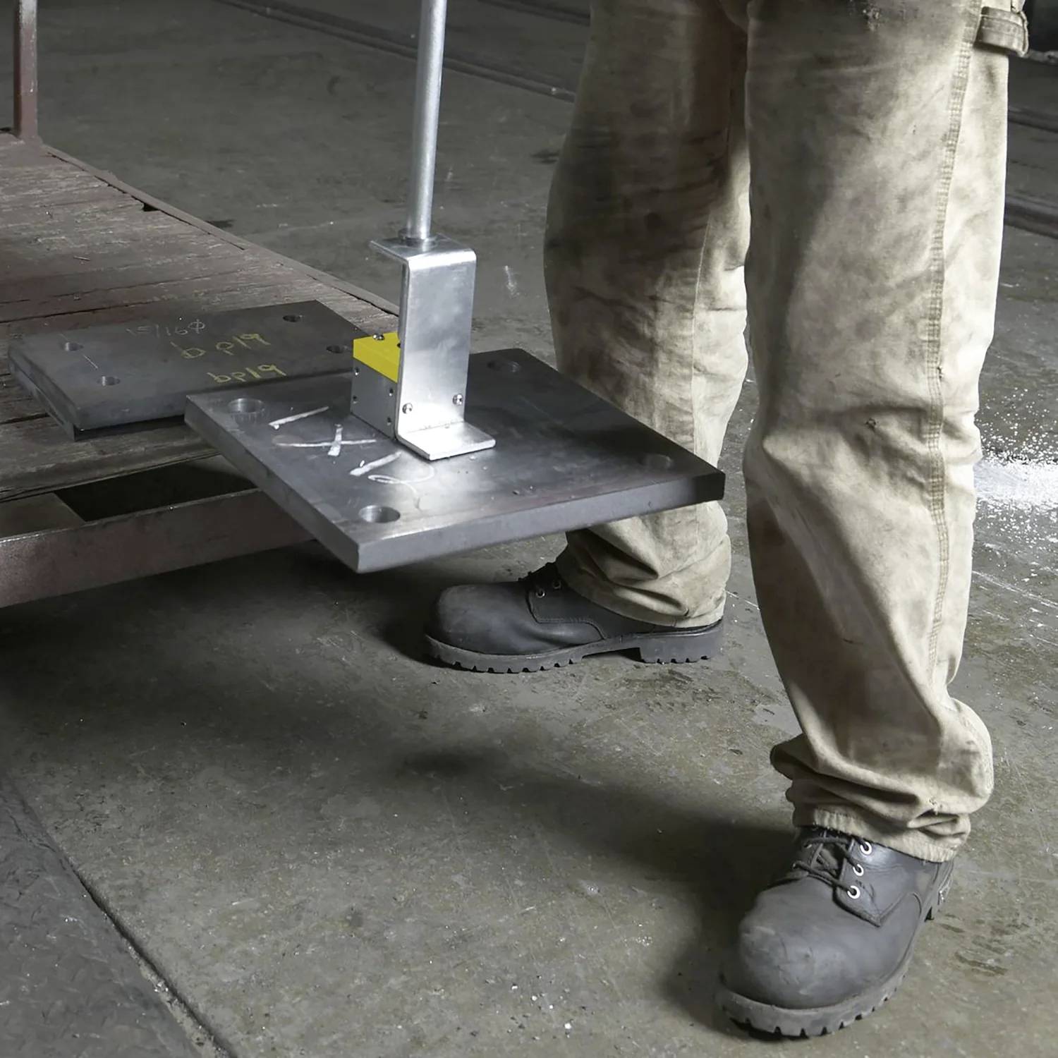 Osoba ubrana w buty ochronne i spodnie robocze podnosi arkusz metalowy za pomocą ręcznego urządzenia dźwigowego w środowisku przemysłowym.