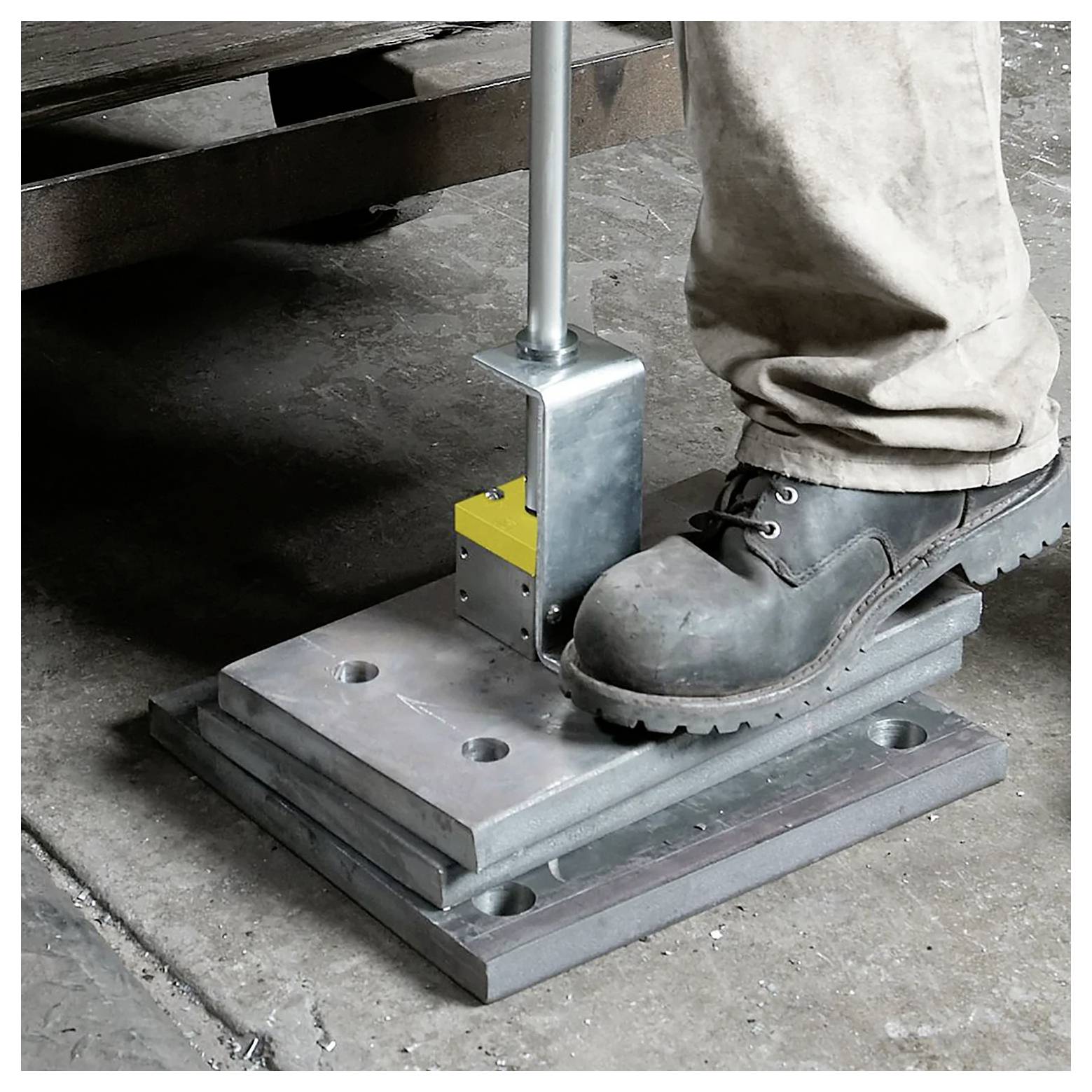 Pracownik obsługujący prasę metalową nogą, ubrany w ochronne buty robocze, na betonowej podłodze w przemysłowym otoczeniu.
