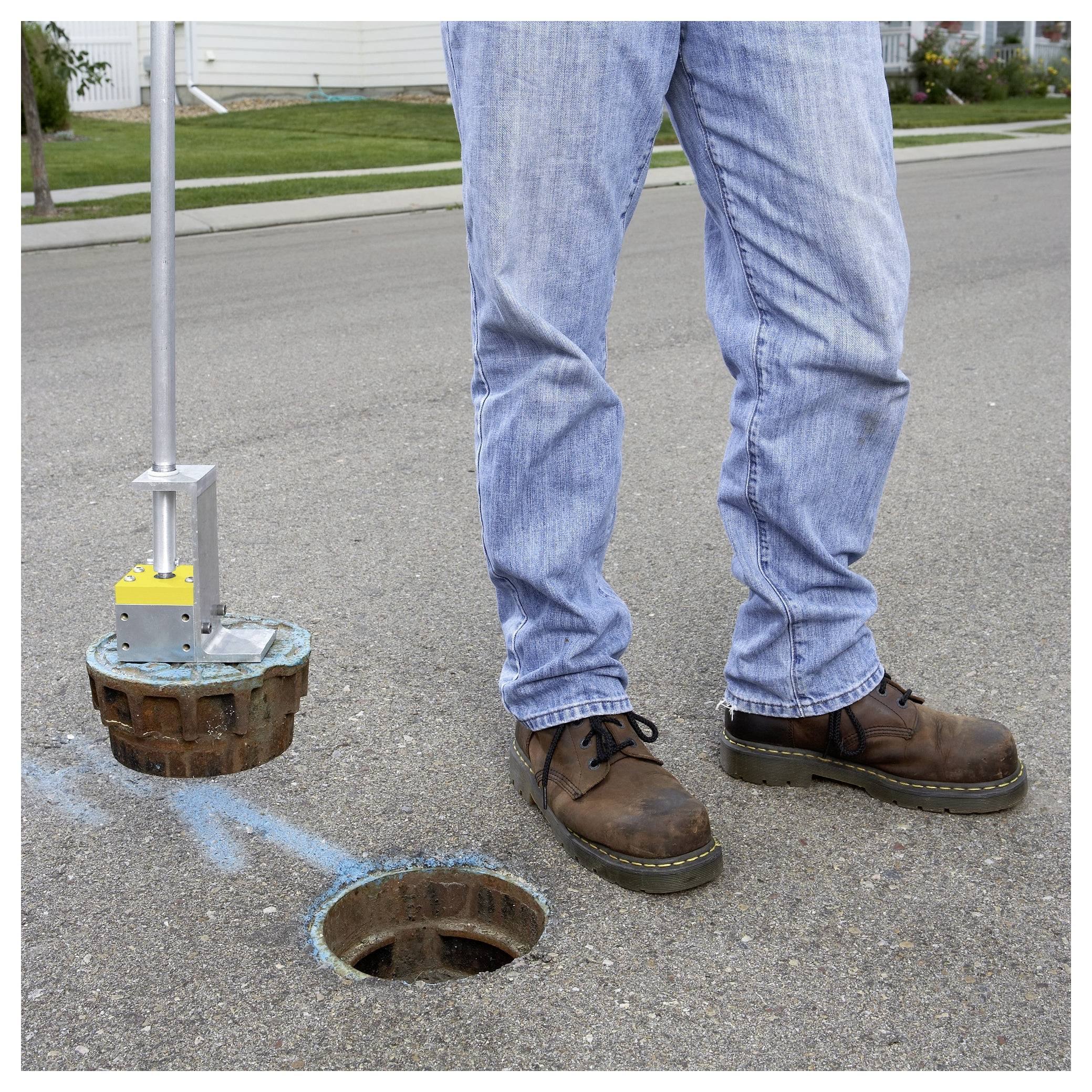 Osoba stoi obok otwartego włazu, trzymając narzędzie przeznaczone do podnoszenia pokrywy, na ulicy mieszkalnej.