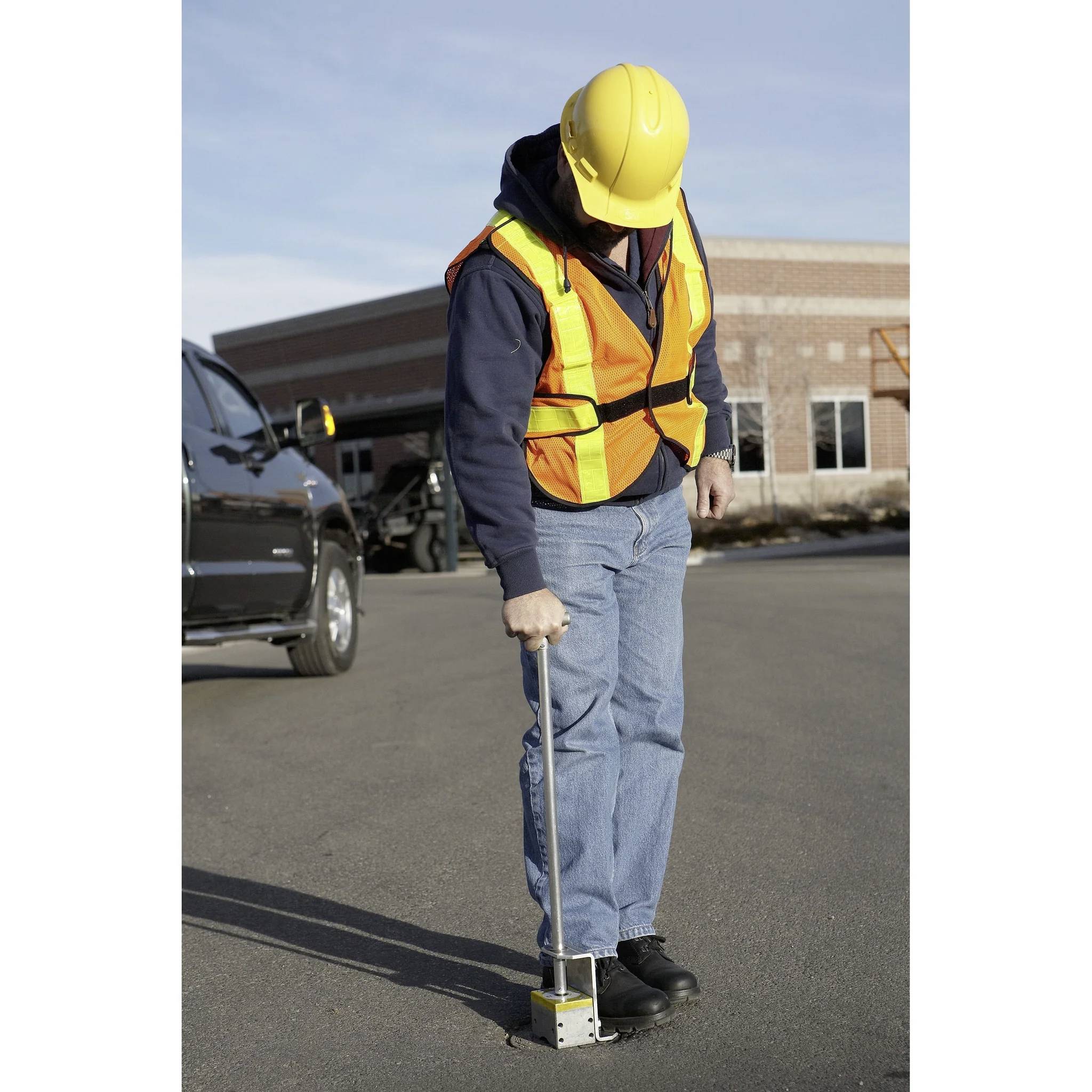 Robotnik budowlany w kamizelce odblaskowej i kasku używa narzędzia do pomiarów gruntu na utwardzonej powierzchni w pobliżu budynku z cegły.