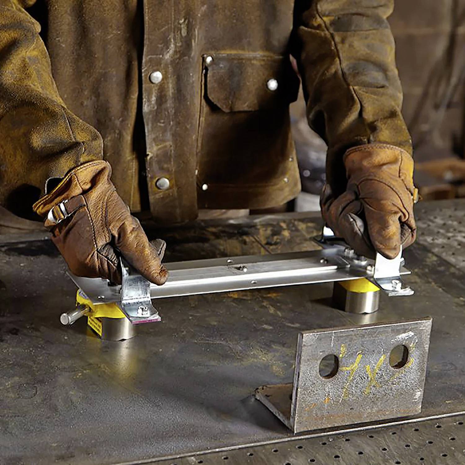 Osoba w rękawiczkach prezentuje mocowanie metalowego elementu do magnetycznego uchwytu na stole warsztatowym, wokół której znajdują się narzędzia i kawałki metalu.