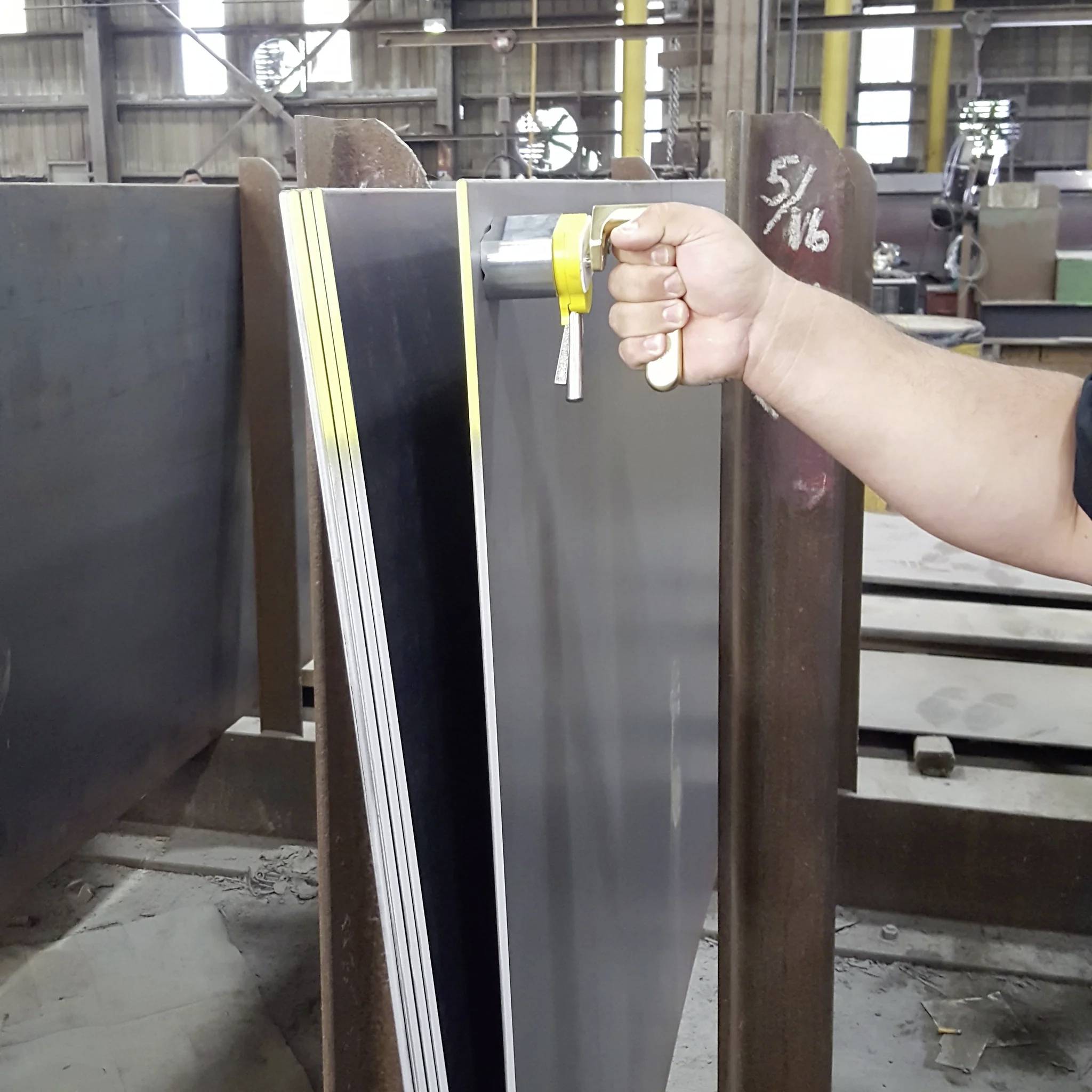 Dłoń trzyma żółty grubościomierz, mierzący kilka ułożonych w stosie arkuszy metalu w warsztacie, oznaczony napisem '5/16'.