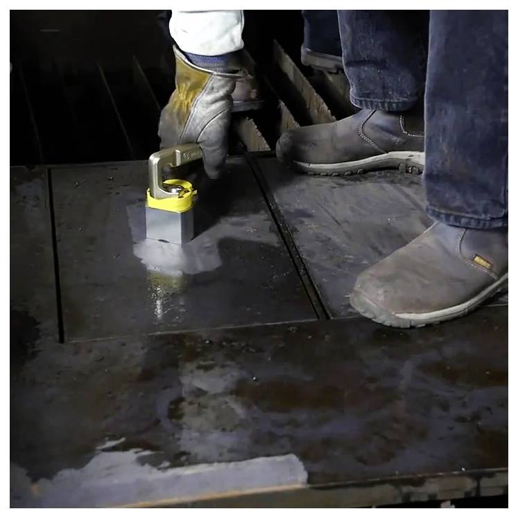 Osoba obsługuje magnetyczne urządzenie transportowe na metalowej powierzchni, nosząc ochronne rękawice i buty, co wskazuje na bezpieczną procedurę przemysłową.