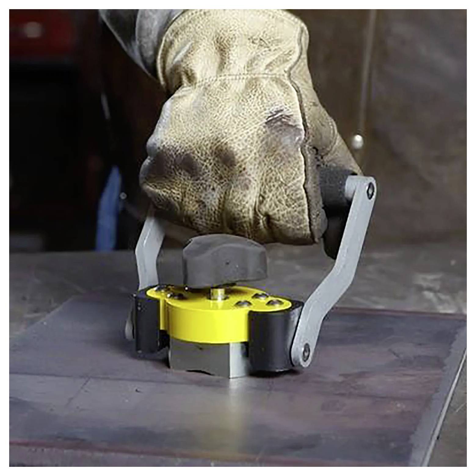 Rękawica trzyma żółty magnetyczny podnośnik na metalowej powierzchni w warsztacie, sugerując obróbkę metalu lub działalność przemysłową.