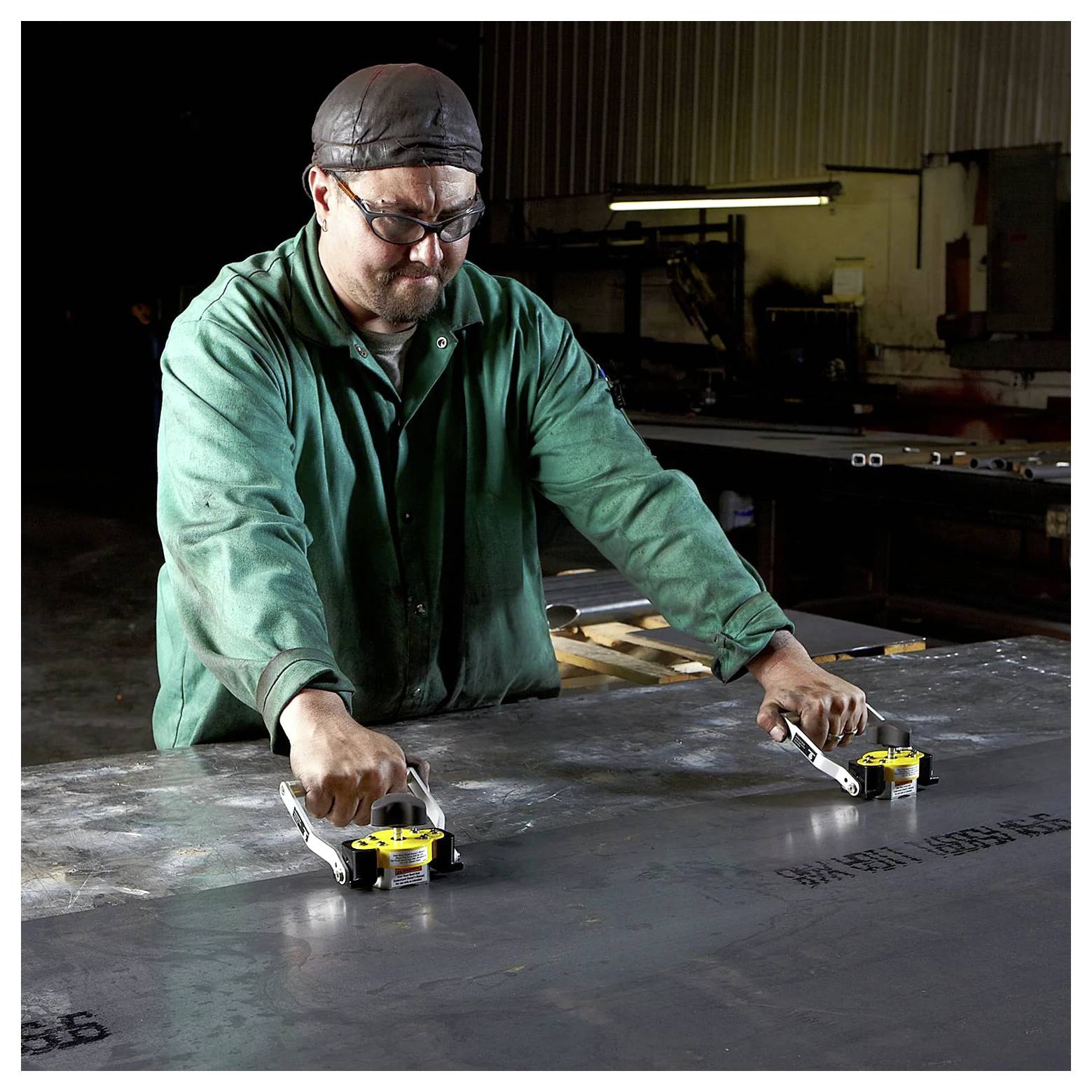 Pracownik w ciemnym warsztacie podnosi arkusz metalu za pomocą dwóch podnośników magnetycznych, ubrany w zieloną kurtkę ochronną, rękawice i czapkę.