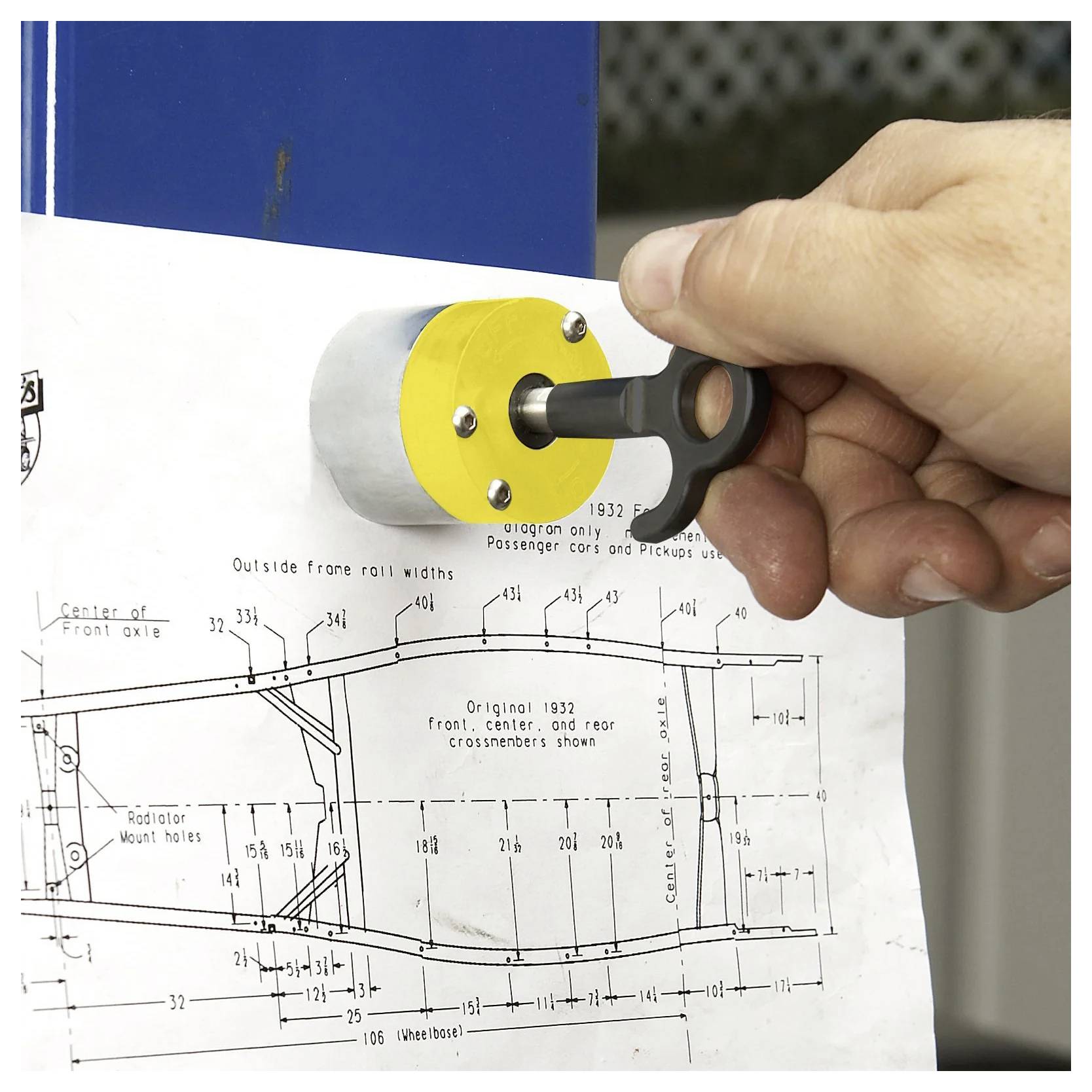 Ręka używa czarnego klucza z żółtą rękojeścią, aby przypiąć techniczny rysunek podwozia pojazdu do niebieskiej metalowej powierzchni.