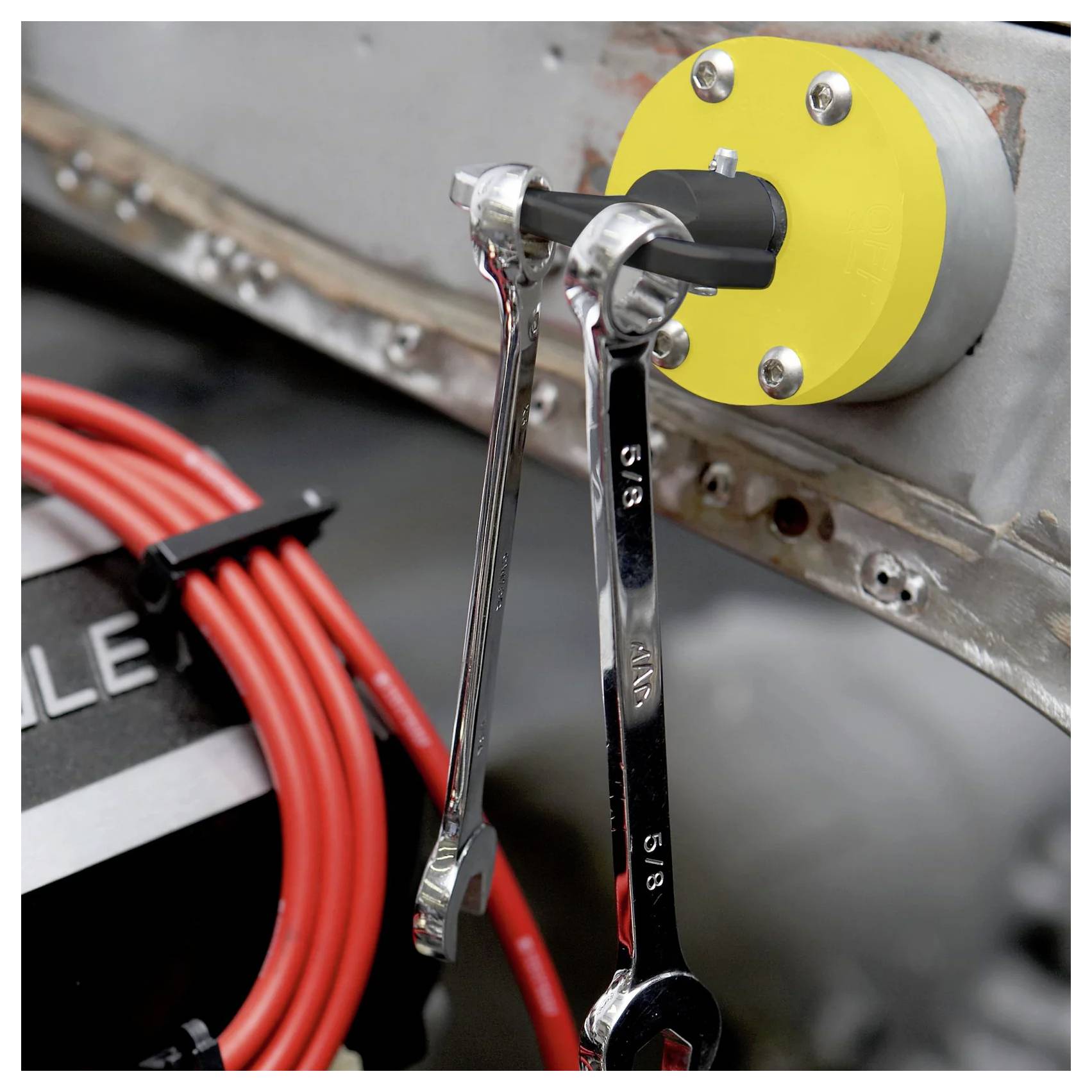 Żółty uchwyt magnetyczny przymocowany do szarej powierzchni, pewnie trzymający dwa metalowe klucze nastawne, z czerwonymi kablami widocznymi w tle.