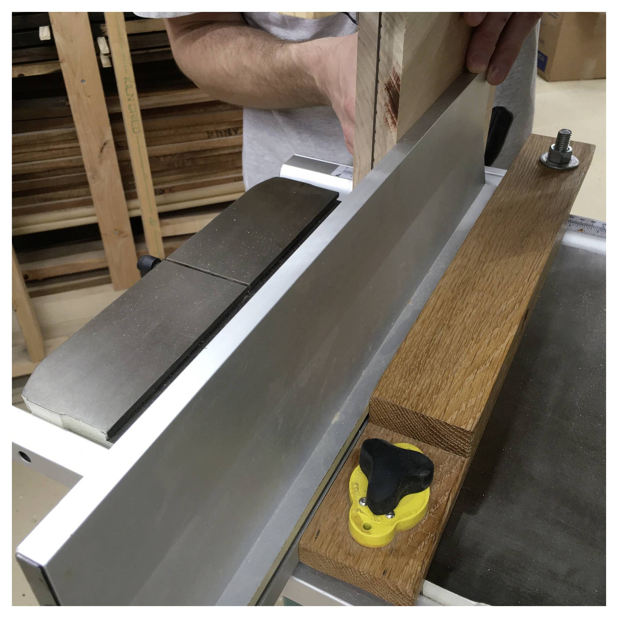 Osoba używa strugarki dolnowrzecionowej, aby wygładzić i wyprostować drewnianą deskę. Widoczne są prowadnica maszyny i ostrze tnące, wokół rozrzucone są wióry drewniane.