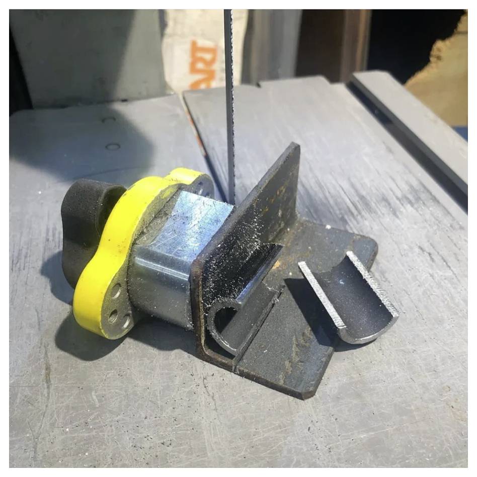 Metalowy zacisk przytrzymujący blok jest przecinany przez pionowe ostrze piły taśmowej w warunkach warsztatowych, podkreślając precyzyjne cięcie.