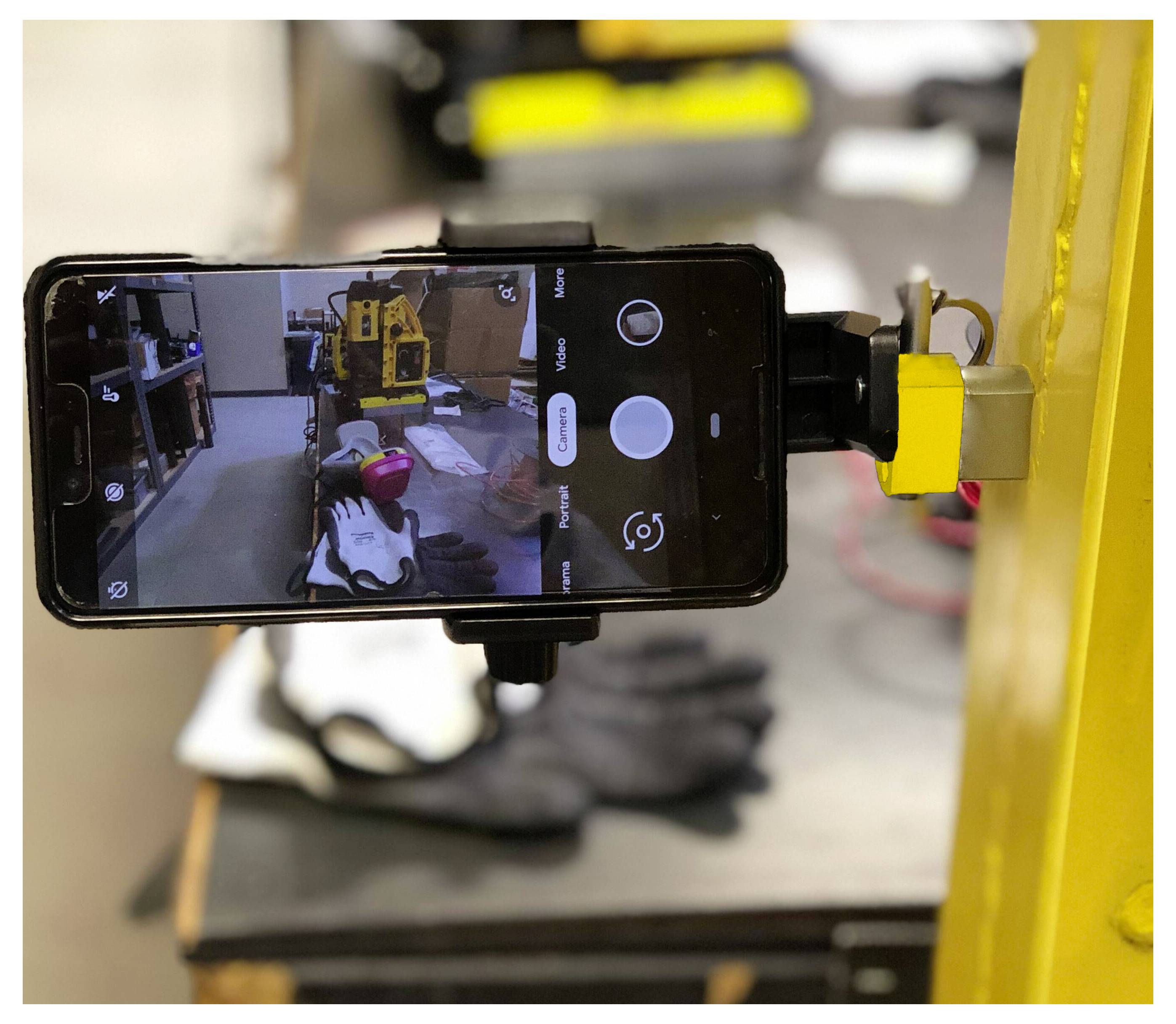Smartfon zamontowany na żółtej maszynie wyświetla widok kamery warsztatu z narzędziami i rękawicą leżącą na stole w tle.