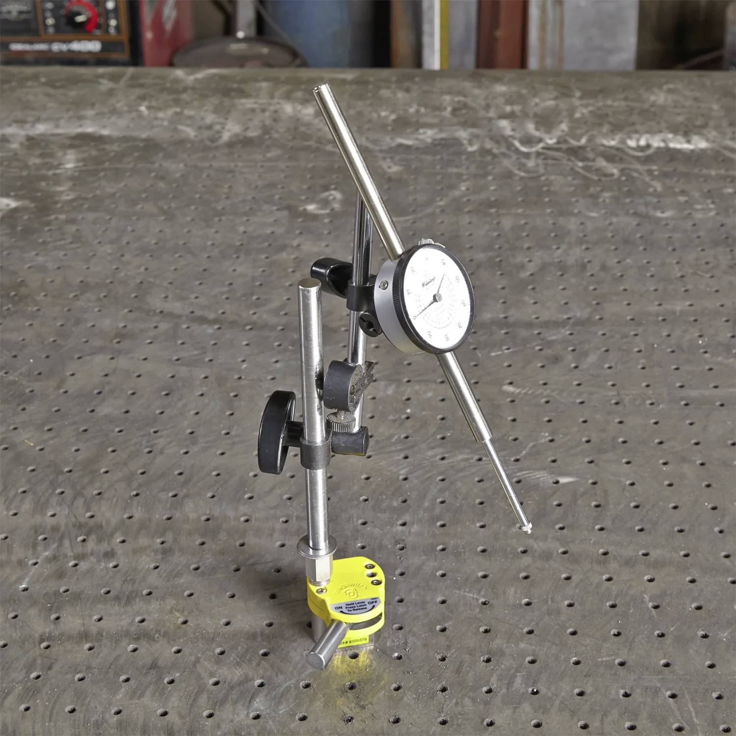 Wskaźnik zegarowy zamontowany na podstawie magnetycznej stoi na perforowanej powierzchni metalowej, używany do precyzyjnych pomiarów w obróbce mechanicznej.