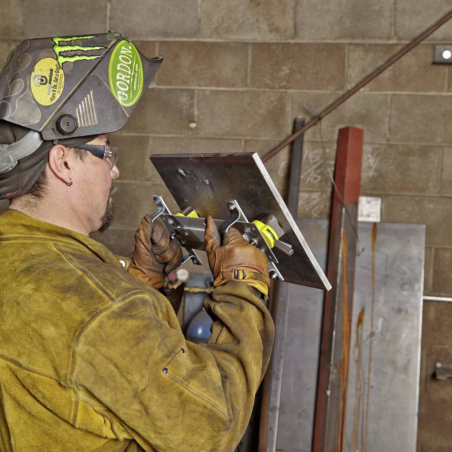 Spawacz w odzieży ochronnej trzyma kawałek metalu w zaciskach, przygotowując się do pracy, na tle ściany warsztatu z narzędziami.