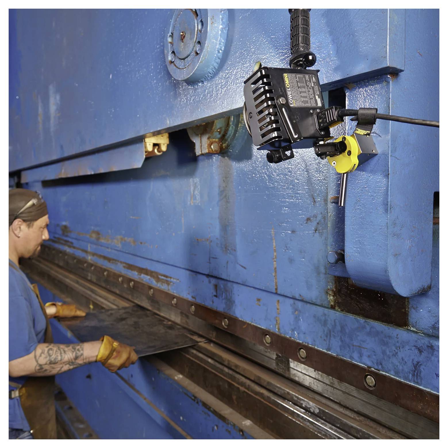 Robotnik fabryczny obsługuje dużą niebieską giętarkę do blachy, prowadząc arkusz metalu do odpowiedniej pozycji, z widocznym sprzętem sterowniczym.