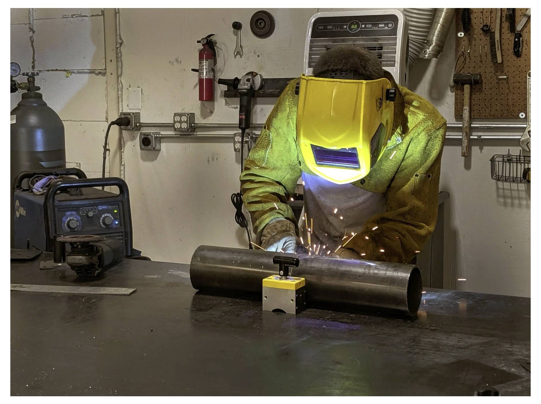 Spawacz w odzieży ochronnej i żółtym kasku pracuje przy metalowej rurze w warsztacie, widoczne są iskry spawalnicze.