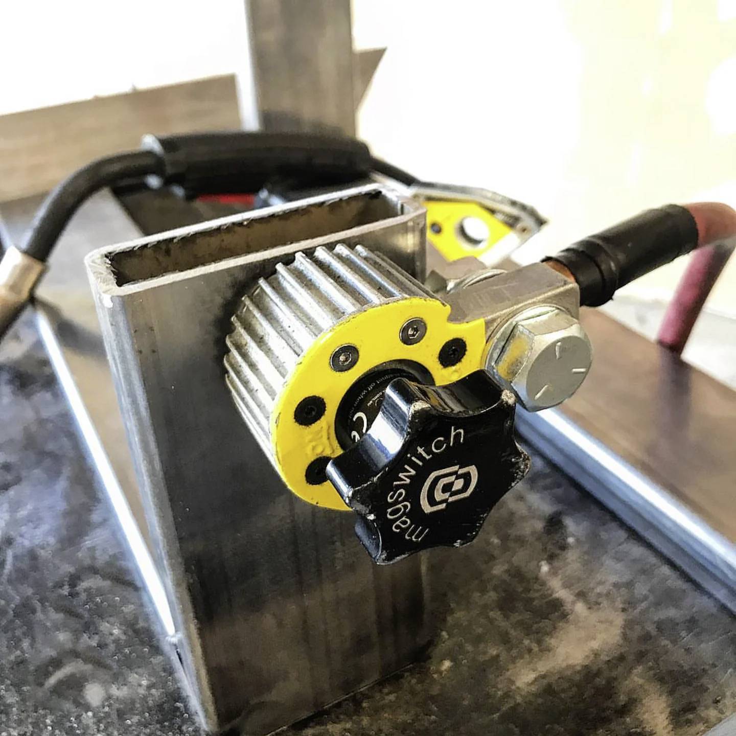 Zbliżenie przestrzeni roboczej warsztatowej z metalową konstrukcją, przedstawiające mechaniczny przełącznik z żółtym kołem zębatym i czarnym pokrętłem oznaczonym symbolami 'O' i 'I'.