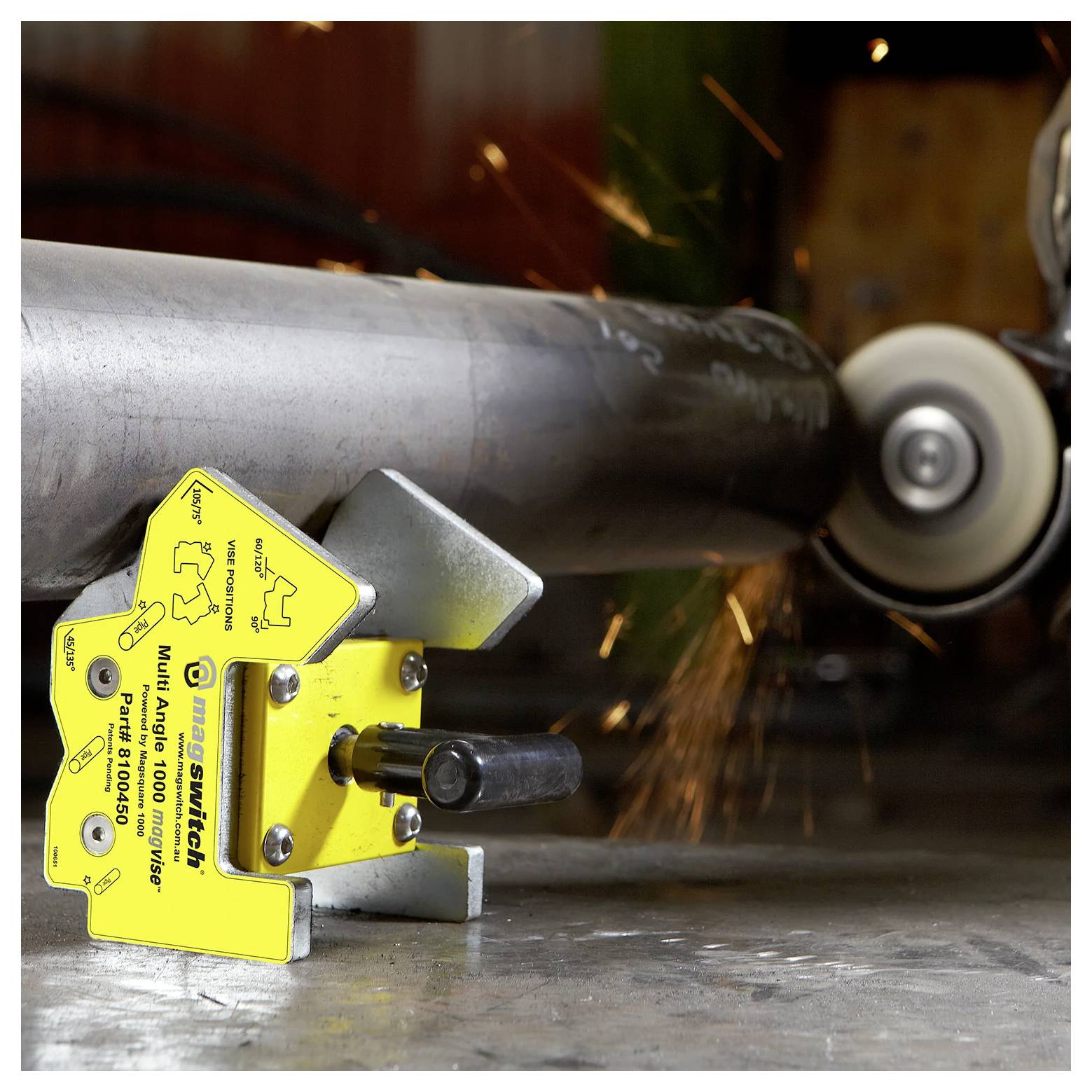 Magnes spawalniczy przytrzymuje metalową rurę, podczas gdy z użytkowanej szlifierki lecą iskry. Magnes jest żółty, oznaczony kątami i instrukcjami bezpieczeństwa.