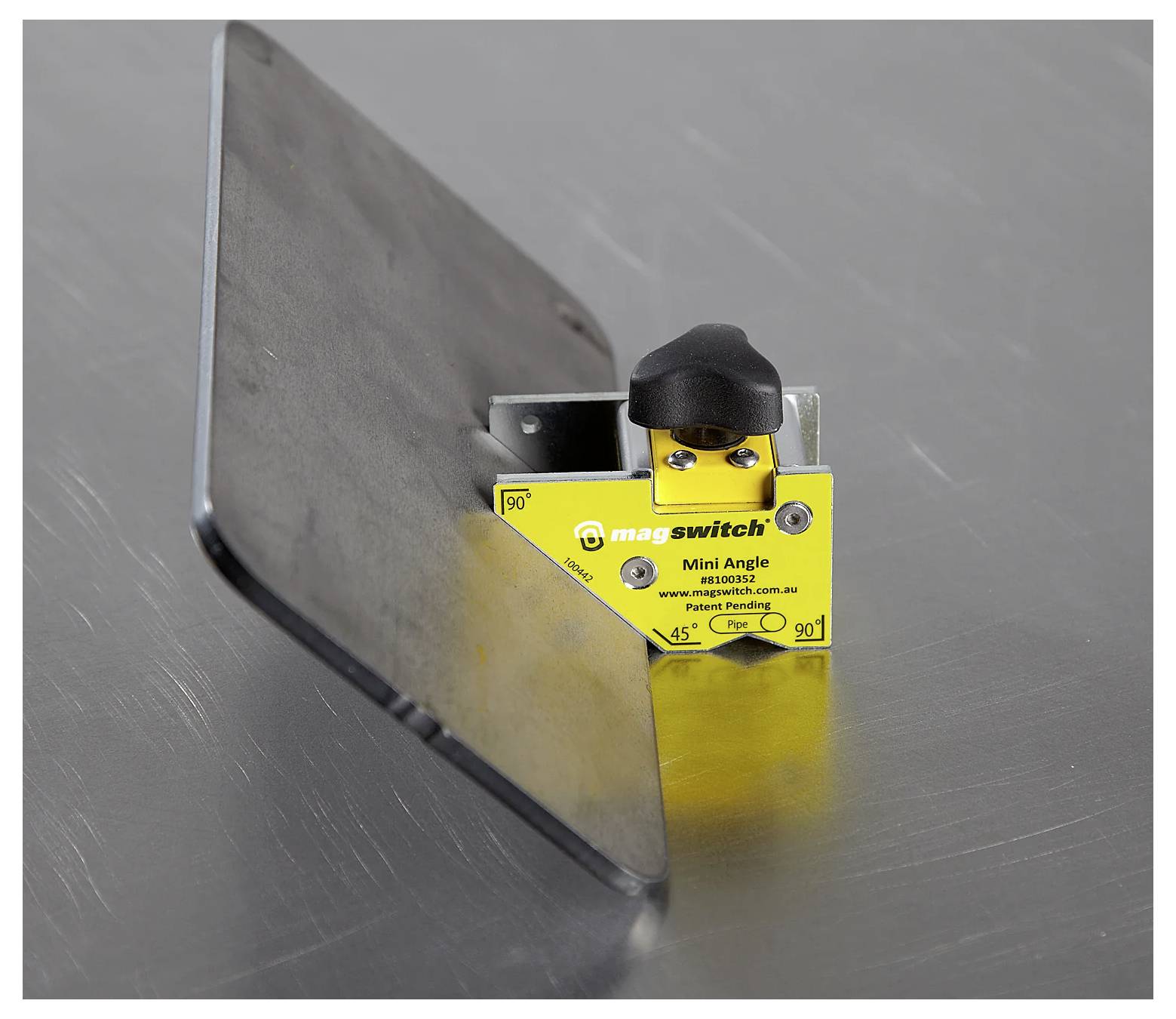 Narzędzie kątowe Magswitch Mini przytrzymujące arkusz metalowy pod kątem 45 stopni na metalowej powierzchni, ilustrujące magnetyczne wsparcie podczas spawania lub pozycjonowania elementów.