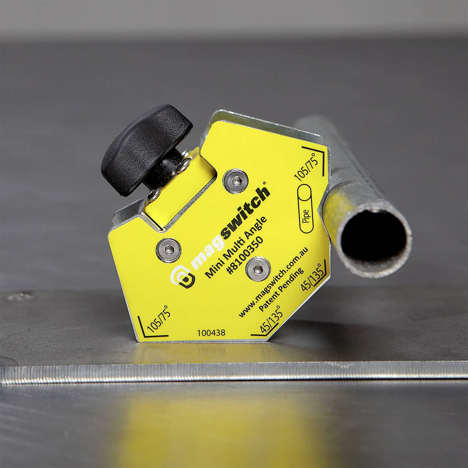 Żółty mini-wielokątny przyrząd Magswitch trzyma metalową rurę na metalowej powierzchni, używany do wyrównywania i mocowania obiektów podczas spawania.