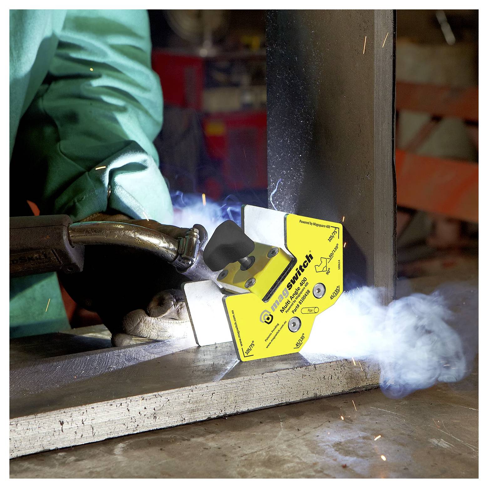 Osoba w odzieży ochronnej spawuje metal, używając żółtego uchwytu magnetycznego do podtrzymania elementów. Dym unosi się nad spawaniem.