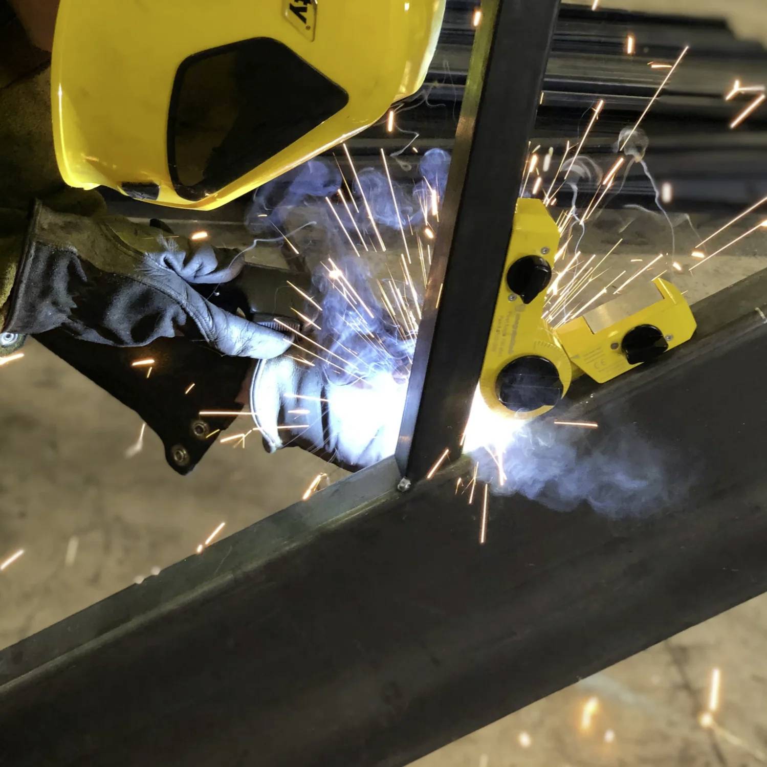 Spawacz w żółtym kasku i rękawicach pracujący, z lecącymi iskrami, trzymający narzędzie spawalnicze na elemencie metalowym, wewnątrz pomieszczenia.