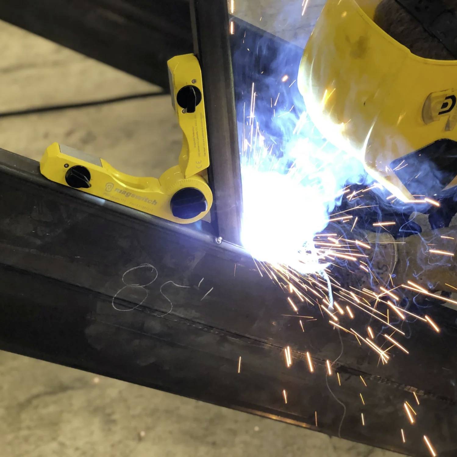 Osoba w żółtym kasku spawalniczym pracuje na metalowej belce, w tle widoczne są jasne iskry spawalnicze i ochronny imadło.