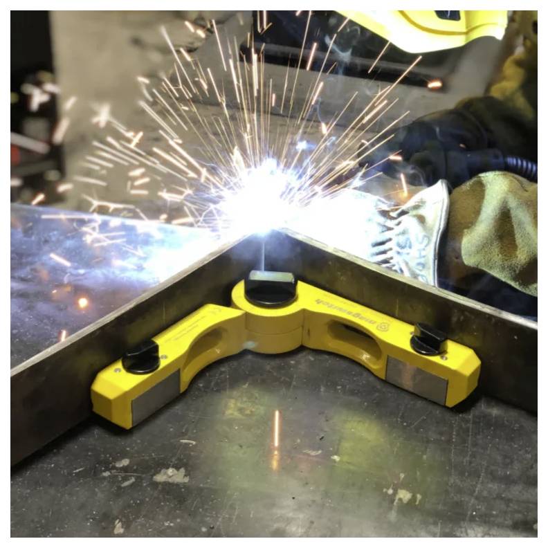 Osoba spawająca przy metalowym złączu, używająca magnetycznego zacisku do spawania narożnego w warsztacie, z lecącymi iskrami podczas procesu spawania.