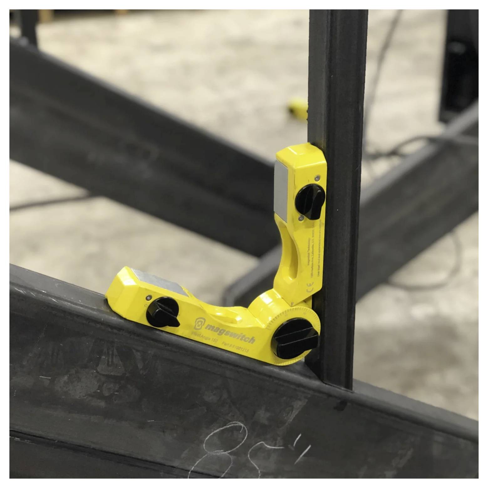 Żółty magnetyczny uchwyt mocujący, zabezpieczający dwie metalowe belki pod kątem prostym w warsztacie, stosowany do precyzyjnego wyrównywania i zastosowań spawalniczych.