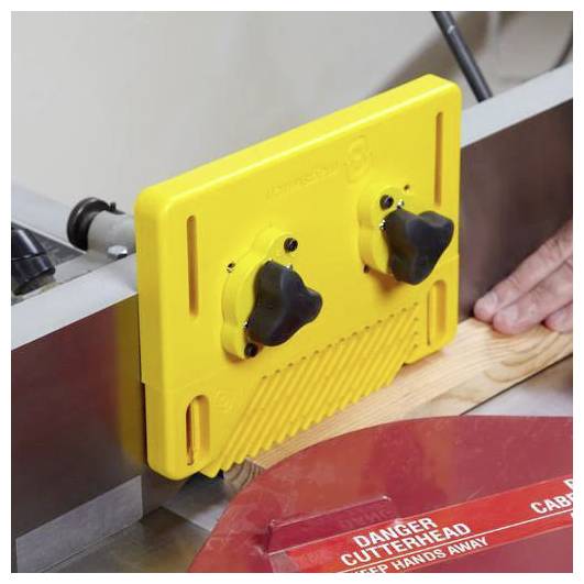 Żółta osłona bezpieczeństwa z dwoma pokrętłami jest zamontowana na pilarce stołowej, a ręka osoby prowadzi kawałek drewna do cięcia.