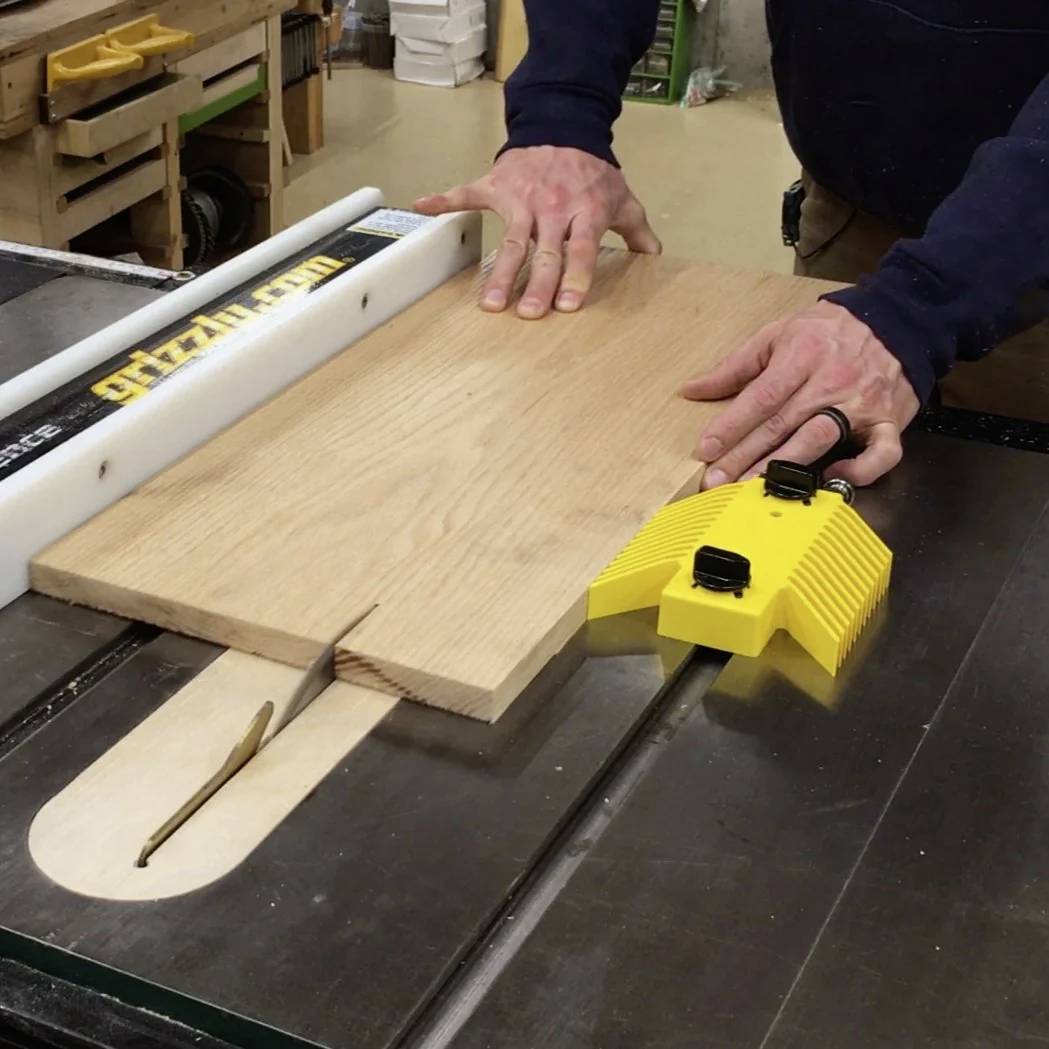 Osoba korzysta z piły tarczowej, prowadząc drewnianą deskę przez ostrze za pomocą narzędzi bezpieczeństwa, prezentując stolarkę w warsztacie.