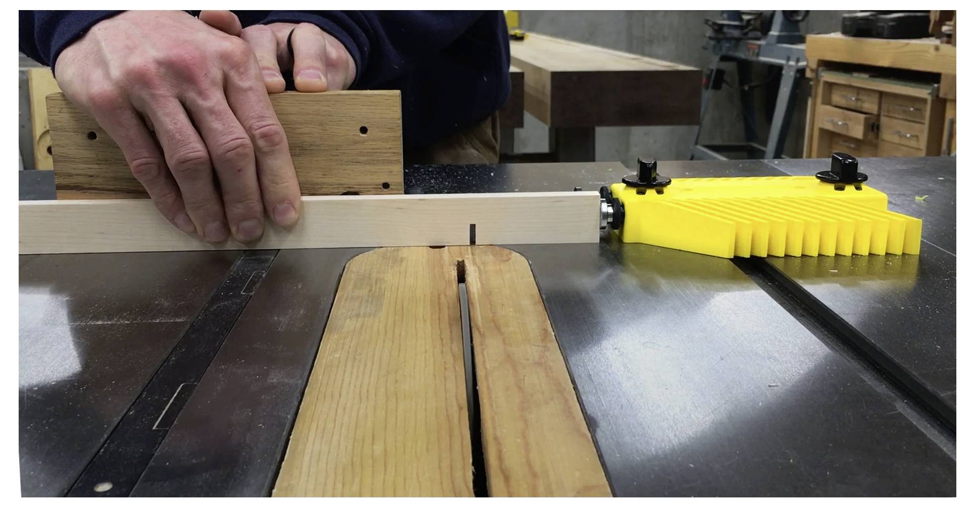 Osoba używa piły stołowej do cięcia kawałka drewna. Ręce prowadzą drewno przez piłę, z widocznymi żółtymi elementami zabezpieczającymi.