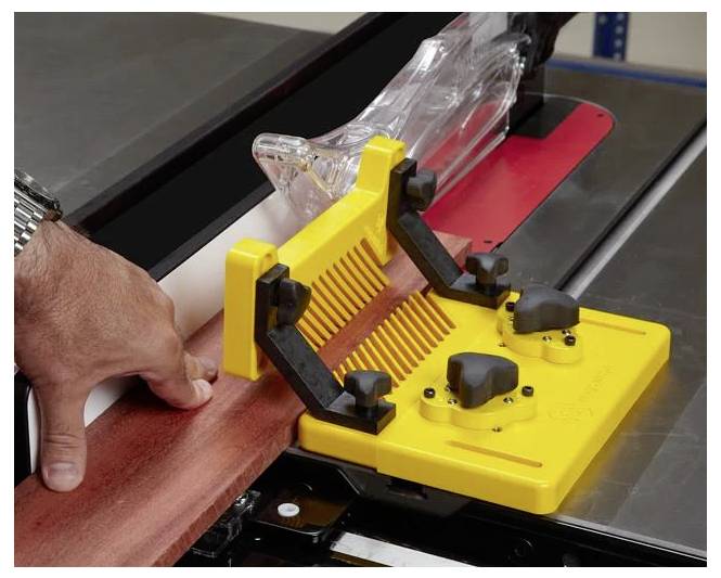 Ręka używająca żółtego popychacza do prowadzenia drewnianej deski przez piłę stołową, demonstrując bezpieczną technikę pracy w stolarstwie.