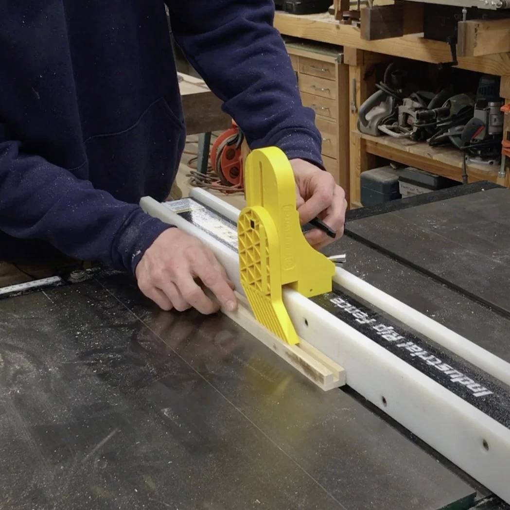 Osoba używa żółtego popychacza do prowadzenia drewna na pilarce stołowej w celu precyzyjnego cięcia w warsztacie.