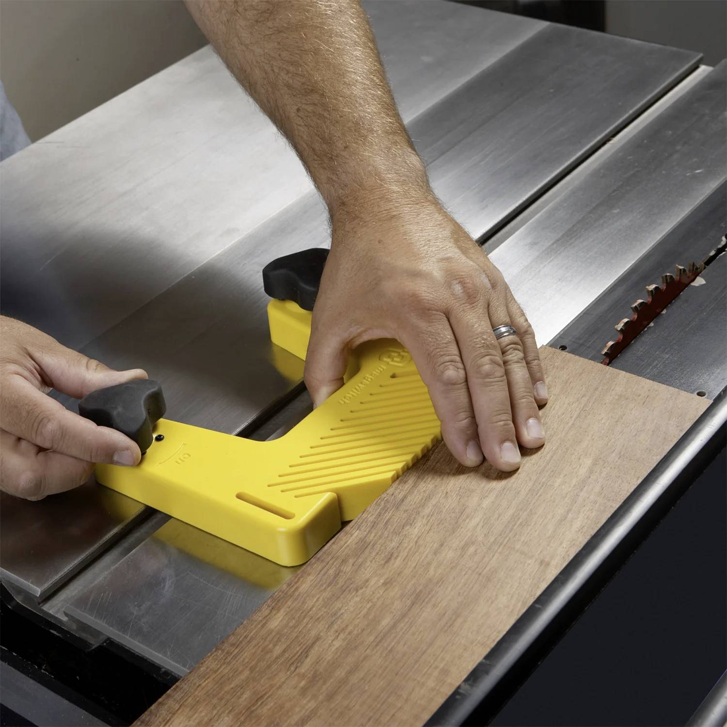 Osoba używa żółtego bloku prowadzącego do bezpiecznego prowadzenia drewnianej deski przez piłę stołową, koncentrując się na bezpieczeństwie dłoni podczas procesu cięcia.