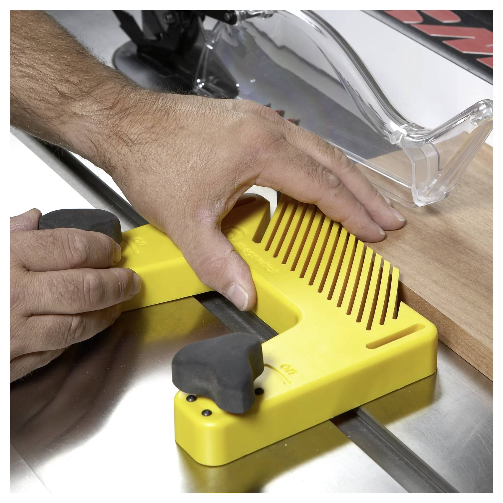 Dłoń osoby reguluje żółte plastikowe urządzenie zabezpieczające na pilarce stołowej, upewniając się, że drewno zostanie pocięte bezpiecznie. Widoczna jest przezroczysta osłona tarczy tnącej.