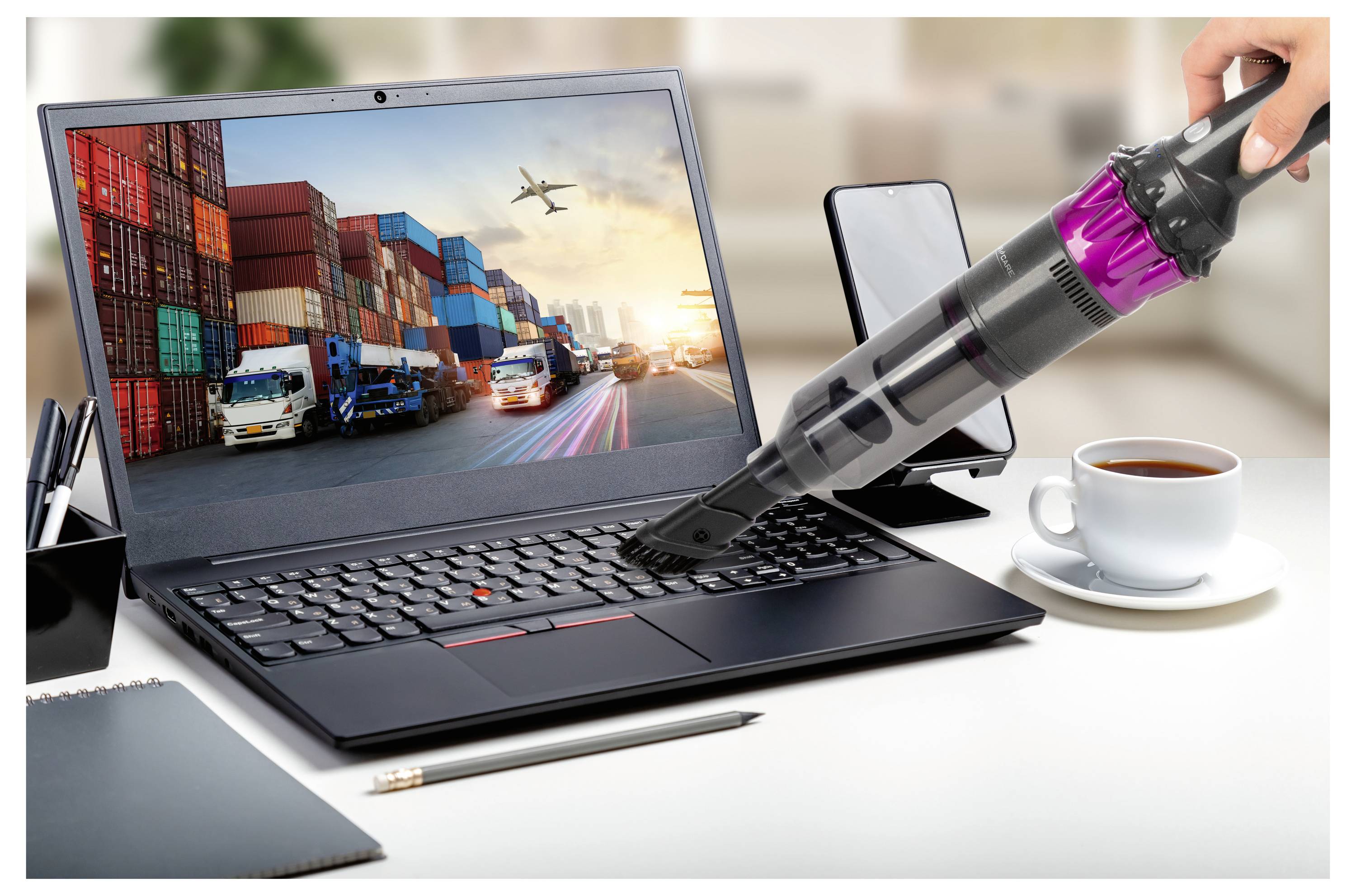 Laptop wyświetla informacje o transporcie ładunku, podczas gdy mini odkurzacz czyści jego klawiaturę. W pobliżu znajdują się smartfon, długopis, notes i kubek kawy.