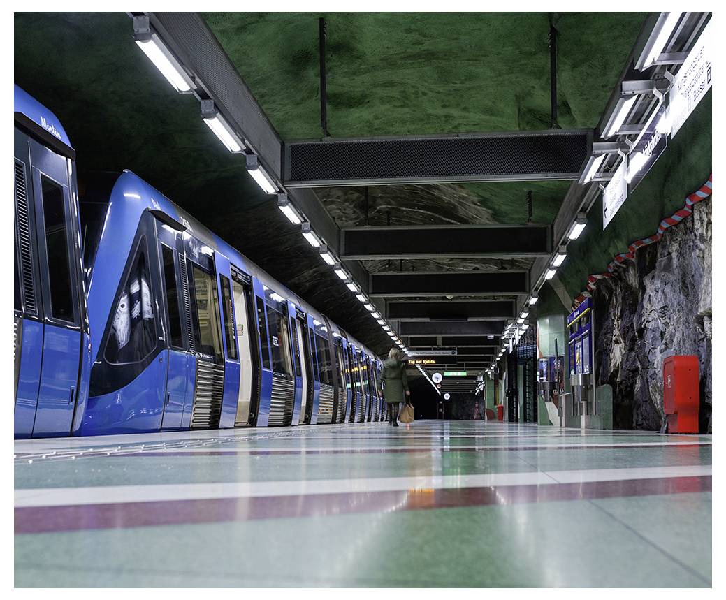 Osoba czekająca na platformie metra z zaparkowanym niebieskim pociągiem w słabo oświetlonej podziemnej stacji, z szorstką, skalistą ścianą w tle.