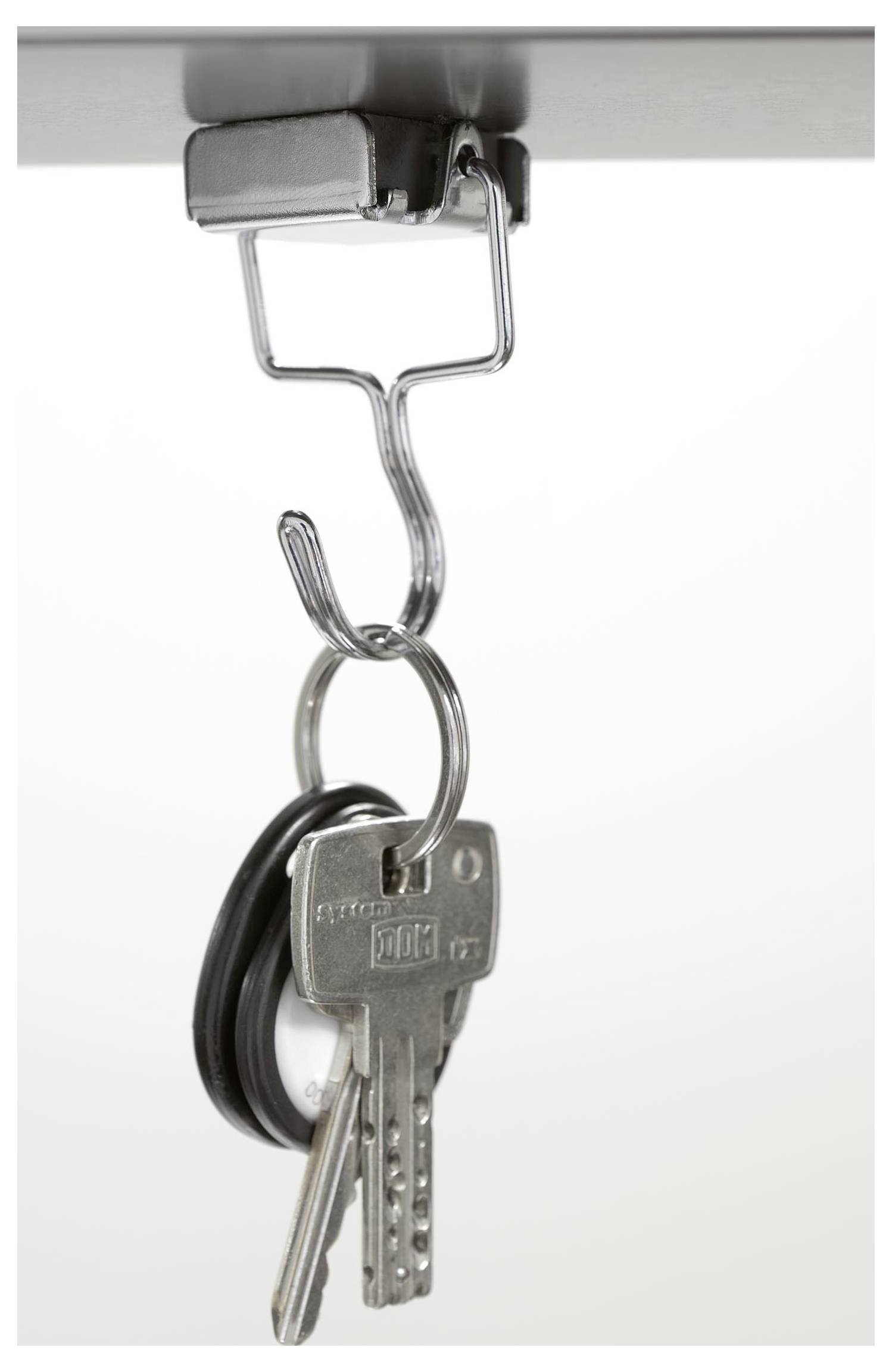 Zestaw kluczy z czarnym brelokiem wisi na metalowym haku pod jakąś powierzchnią, prawdopodobnie stołem lub biurkiem, co sugeruje zorganizowane przechowywanie.